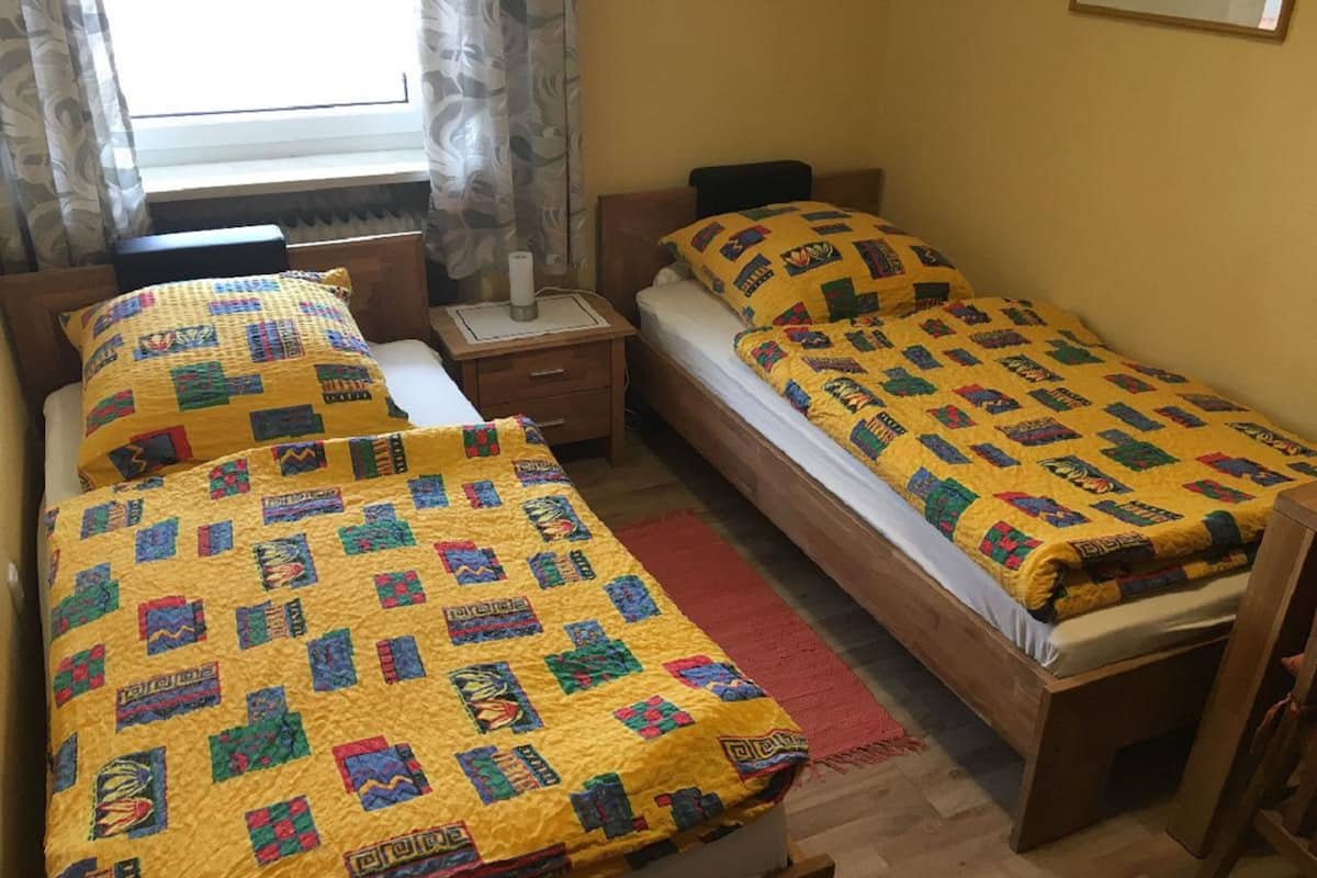 Una habitación con dos camas, cada una con colcha amarilla de motivos coloridos. Entre las camas hay una mesita de noche con lámpara, y una ventana con cortinas.