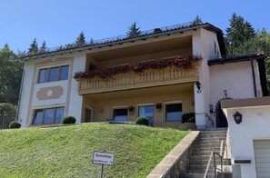 Een tweelaags gebouw met een balkon versierd met rode bloemen, een bordje dat aangeeft dat het een pension is, en een trap die naar de ingang leidt.