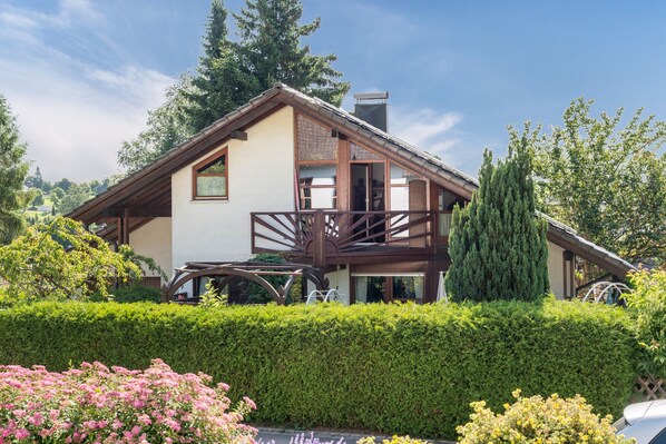 A two-story house with a balcony, surrounded by greenery and a hedge.