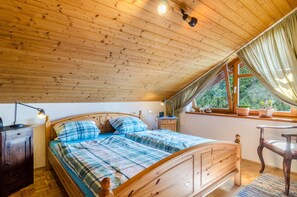 A wooden ceiling, a bed with a plaid bedspread, a nightstand, a chair, and a window with curtains.