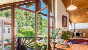A wooden deck with a view of a forest, a dining area with a table and chairs, and a kitchen with appliances. - Holiday Apartment “Rose Garden” near Lake Constance with Wi-Fi & Balcony (Heiligenberg)