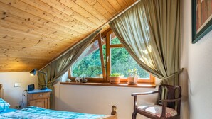 A bedroom with a wooden ceiling, a bed with a blue and white checkered bedspread, a wooden nightstand, a desk with a lamp, a chair, and a window with curtains. - Holiday Apartment “Rose Garden” near Lake Constance with Wi-Fi & Balcony (Heiligenberg)