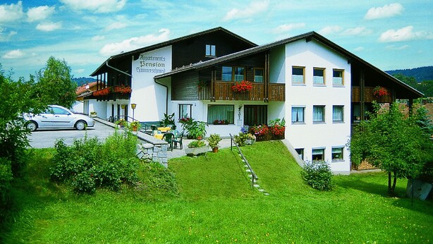 A two-story building with a balcony and flower boxes, surrounded by greenery.