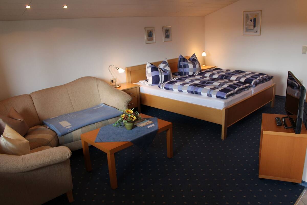 A hotel room with a bed, a sofa, a small table with a vase of flowers, a desk with a mirror, and a television.