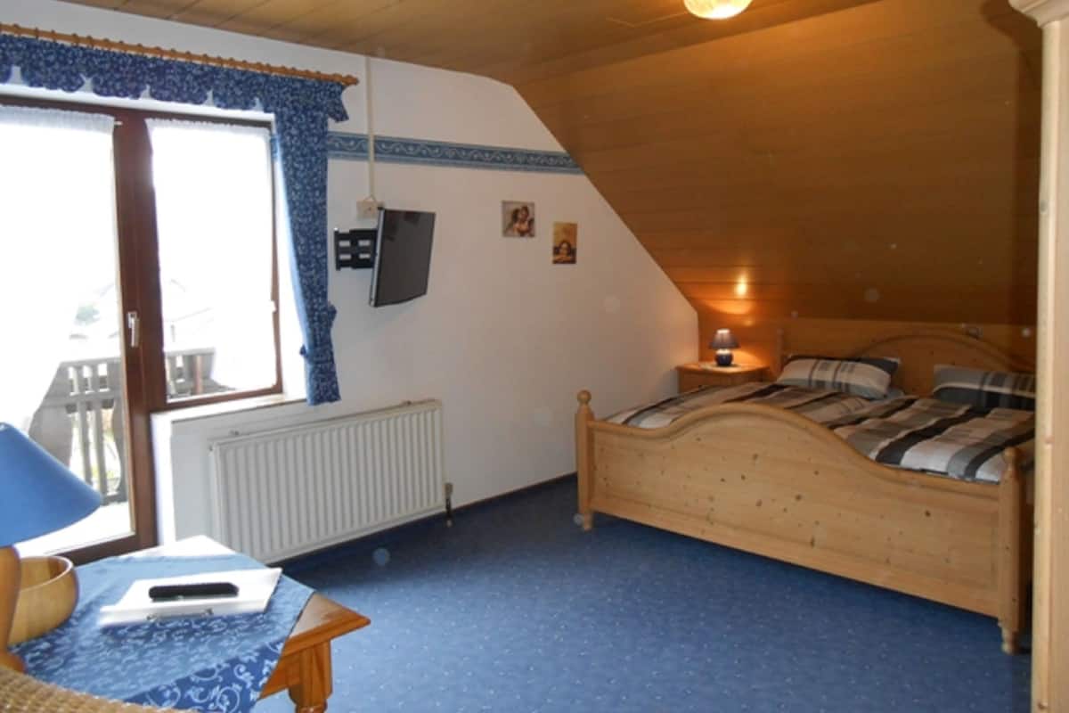 A bedroom with a wooden bed, a table with a lamp, a TV on the wall, and a window with blue curtains.
