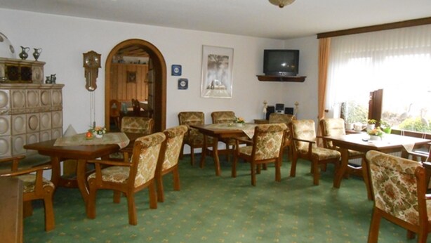 A dining area with a green patterned carpet, wooden chairs, a wooden table, a television mounted on the wall, and a window with curtains. - Pension Sonnenblick (Winterberg)