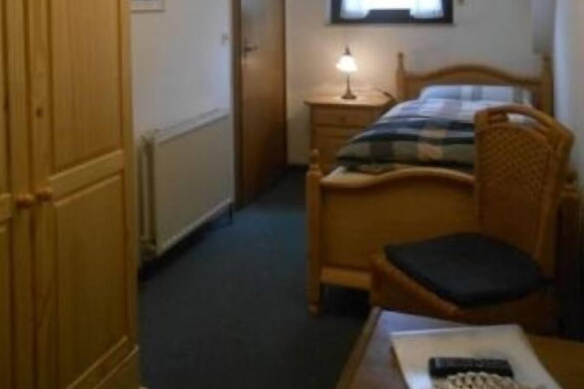 A bedroom with a wooden wardrobe, a bed with a headboard, a bedside table with a lamp, and a window with blinds.