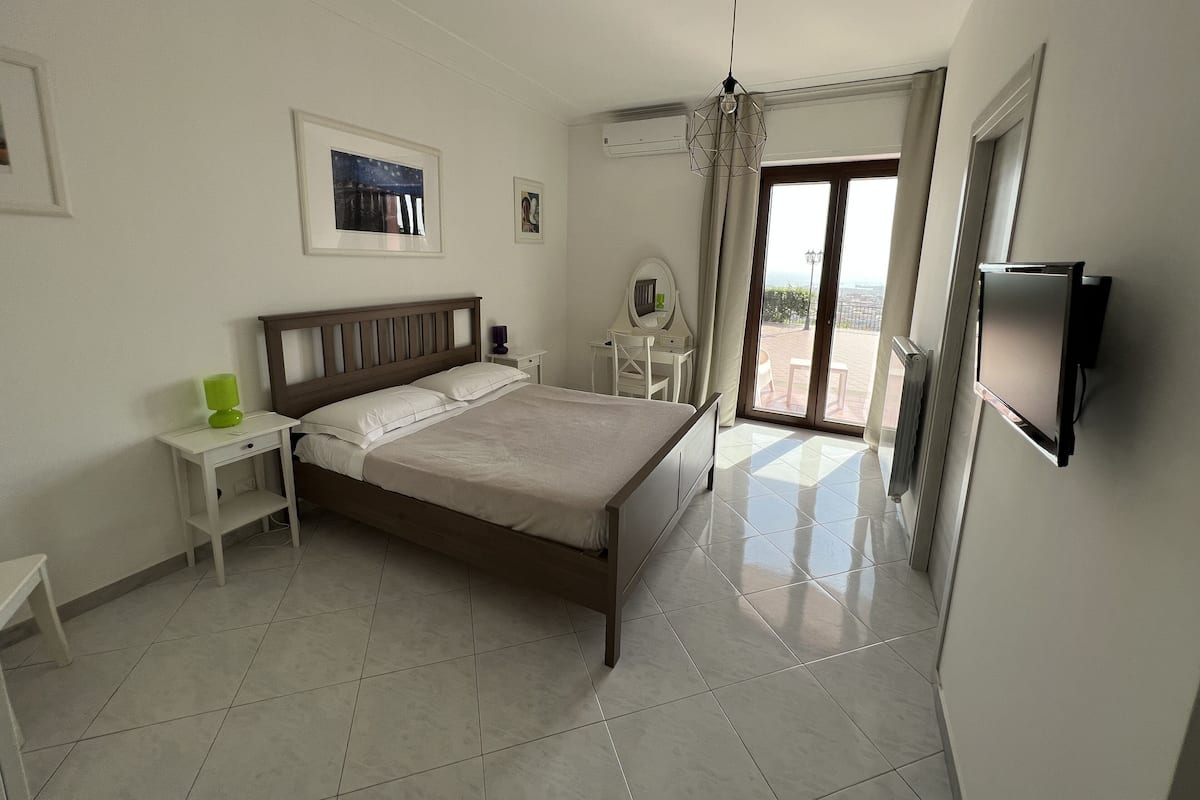 A bedroom with a bed, a desk, a chair, a TV, and a balcony with a view.