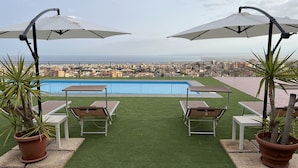 A rooftop pool area with two umbrellas, lounge chairs, and potted plants.