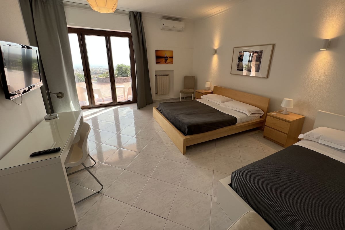 A modern hotel room with a bed, bedside tables, a desk, a TV, and a large window with curtains.