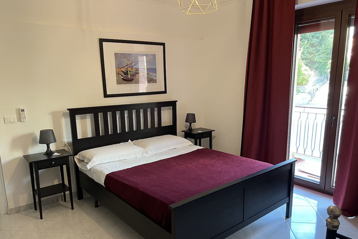 A neatly made bed with a dark headboard, two bedside tables with lamps, a framed artwork above the bed, and a view of the outside through a sliding door.