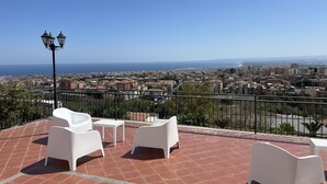 A terrace with white chairs and a table, overlooking a cityscape with buildings and the sea.