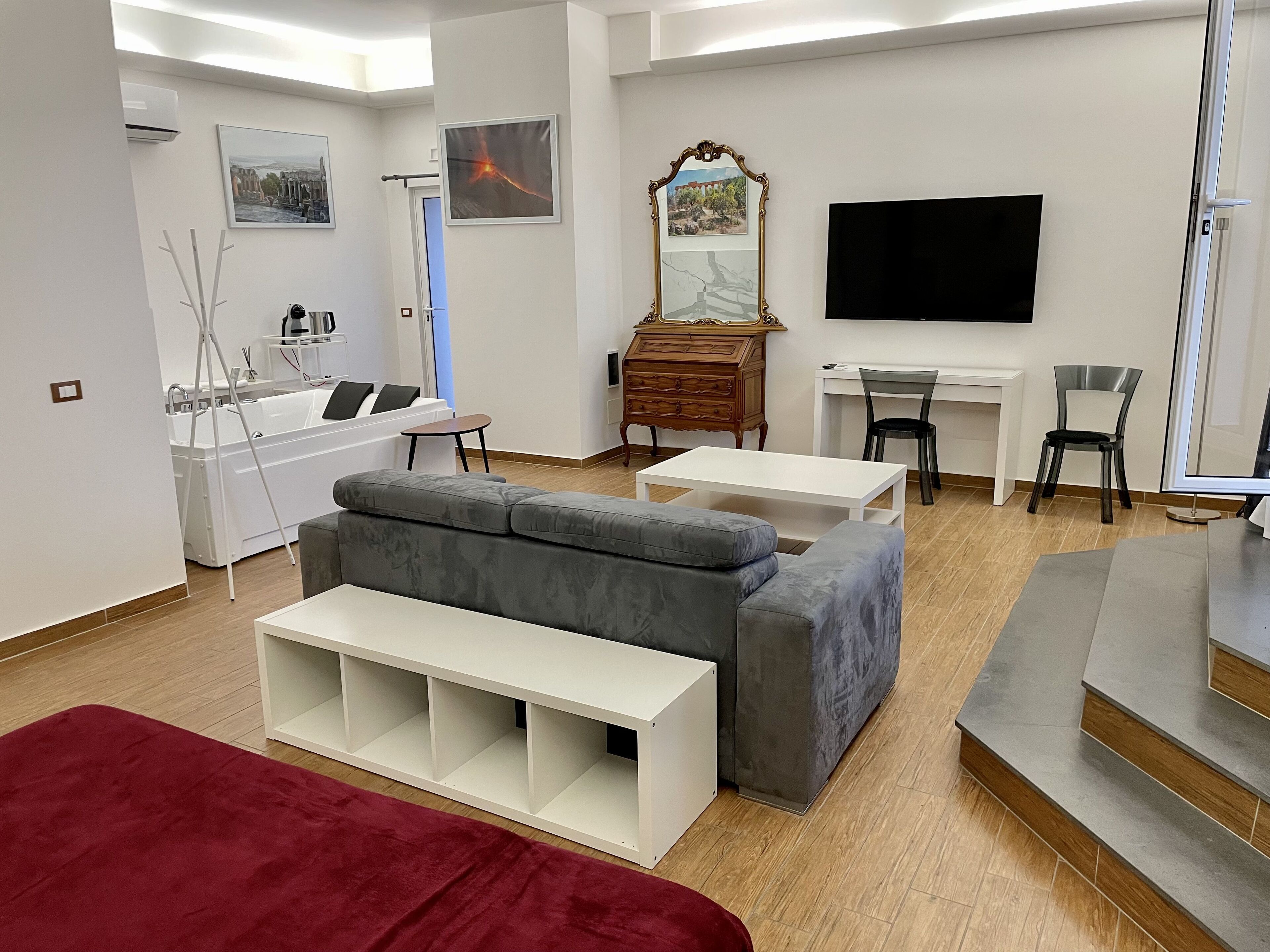 A modern living room with a grey sofa, a red ottoman, a white coffee table, a wooden dresser, a TV, and a desk with two chairs.
