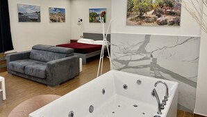 A modern bedroom with a bed, a gray sofa, a white bathtub, and three landscape paintings on the wall.