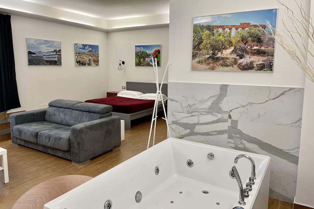 A modern bedroom with a bed, a gray sofa, a white bathtub, and three landscape paintings on the wall.
