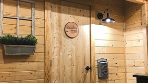 Interno di una baita di legno con una porta su cui è scritto 'L'avventura ti aspetta', una finestra con una pianta, una lampada a parete e una cassetta delle lettere.