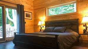 Camera da letto con pannelli in legno, un letto, due lampade da comodino, un quadro incorniciato e una finestra vista alberi.