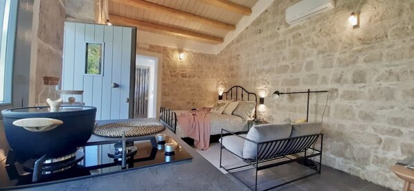 A cozy room with a stone wall, a bed with pillows, a sofa, a table with a bowl, and a wall-mounted lamp.
