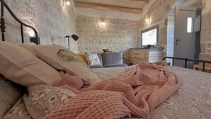 A bedroom with a stone wall, a bed with a pink blanket, a nightstand, a lamp, and a window. - Cottage Myrtle (Giarratana)