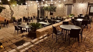 An outdoor patio area with stone walls, multiple tables and chairs, string lights, and potted plants. - Cottage Myrtle (Giarratana)