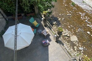 Un patio al aire libre con un parasol blanco, una mesa redonda con un cojín colorido y una mesita con un plato y una copa.