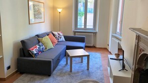 A living room with a sofa, a small table, a lamp, a fireplace, and a window with a view of the street. - Bridge Apartment Argegno (Argegno)
