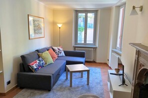 Ein Wohnzimmer mit einer Couch, einem kleinen Tisch, einer Lampe, einem Kamin und einem Fenster mit Blick auf die Straße.