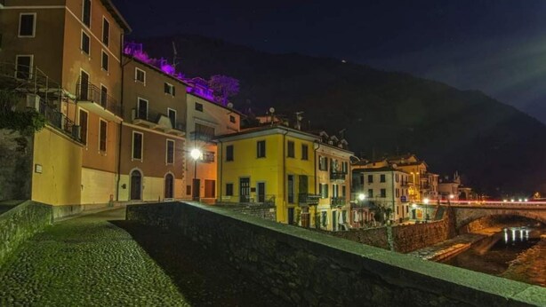 A nighttime view of a coastal town with buildings along a waterfront, a stone wall, and a bridge. - Bridge Apartment Argegno (Argegno)