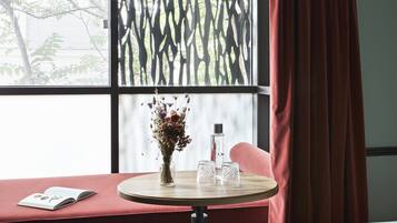 A room with a large window, a round table with a vase of flowers, a book, and a bottle.
