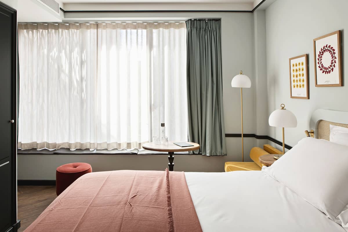 A hotel room with a bed, a round table with a lamp, a chair, and a small stool.
