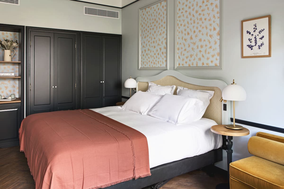 A neatly made bed with a white and rust-colored blanket, a bedside table with a lamp, and a small sofa.