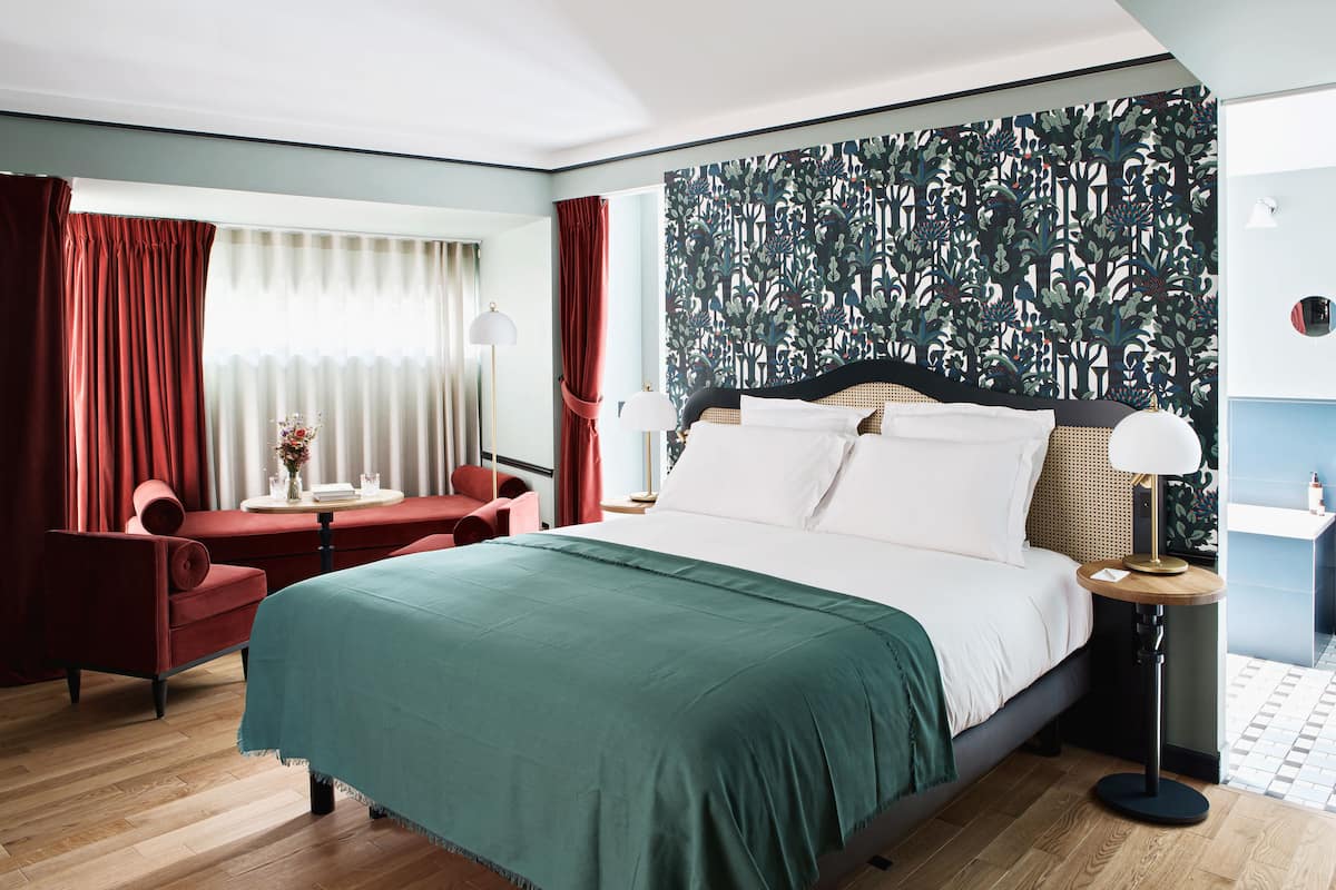 A hotel room with a large bed, a red armchair, a small table with a vase, and a patterned wallpaper on the wall.