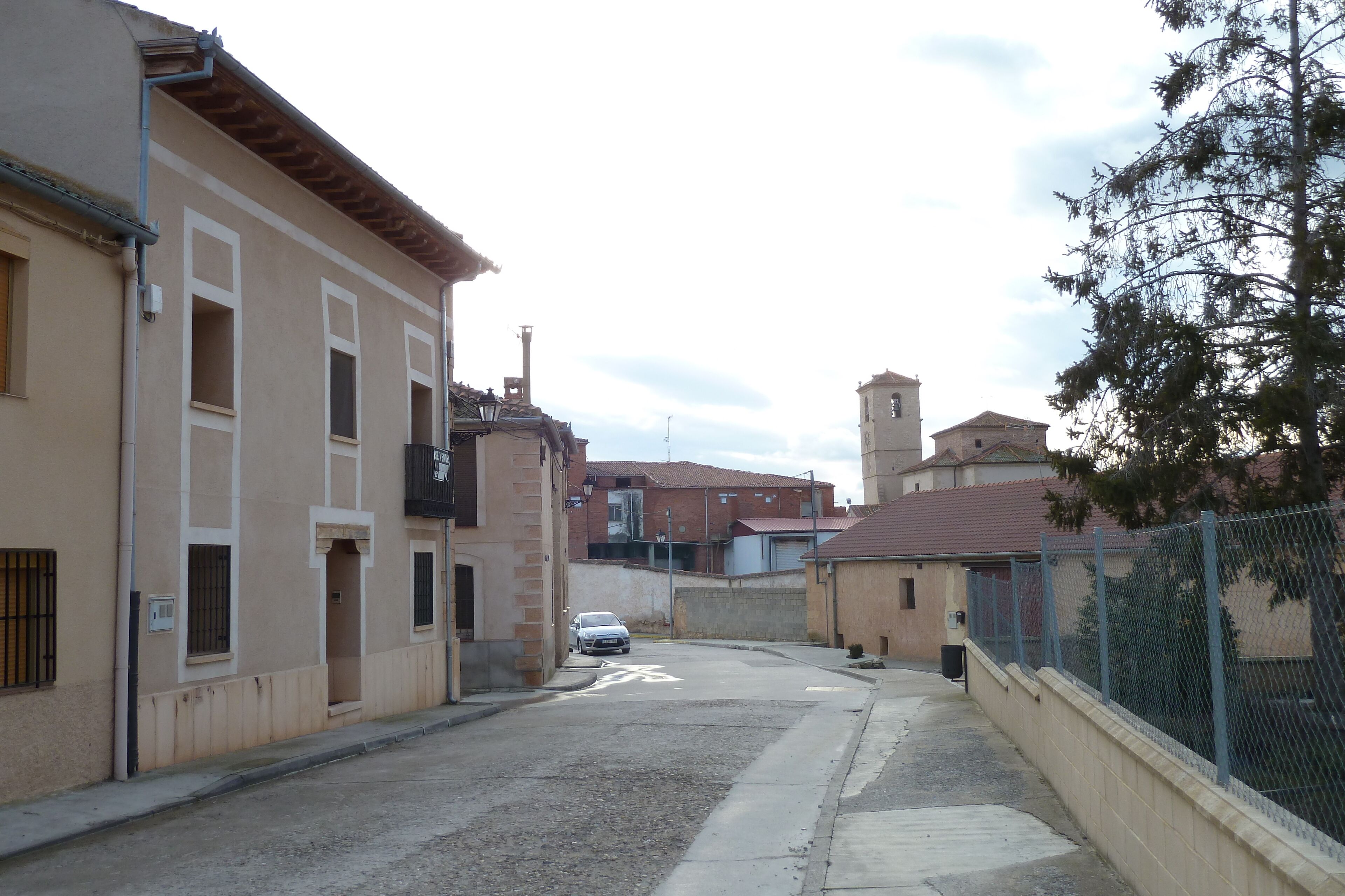 A narrow street with buildings on both sides, a white car parked on the left, and a tall tower in the background.