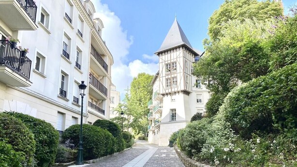 Un sentier pavé de pierres mène à un bâtiment historique doté d’une tour, qui est entouré d’une végétation luxuriante et de bâtiments modernes.