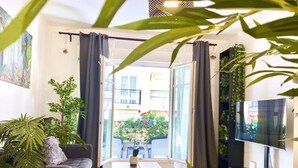 Un salon moderne comprenant un canapé, une table basse, un téléviseur et de grandes fenêtres donnant sur un balcon agrémenté de plantes et offrant une vue sur les bâtiments.