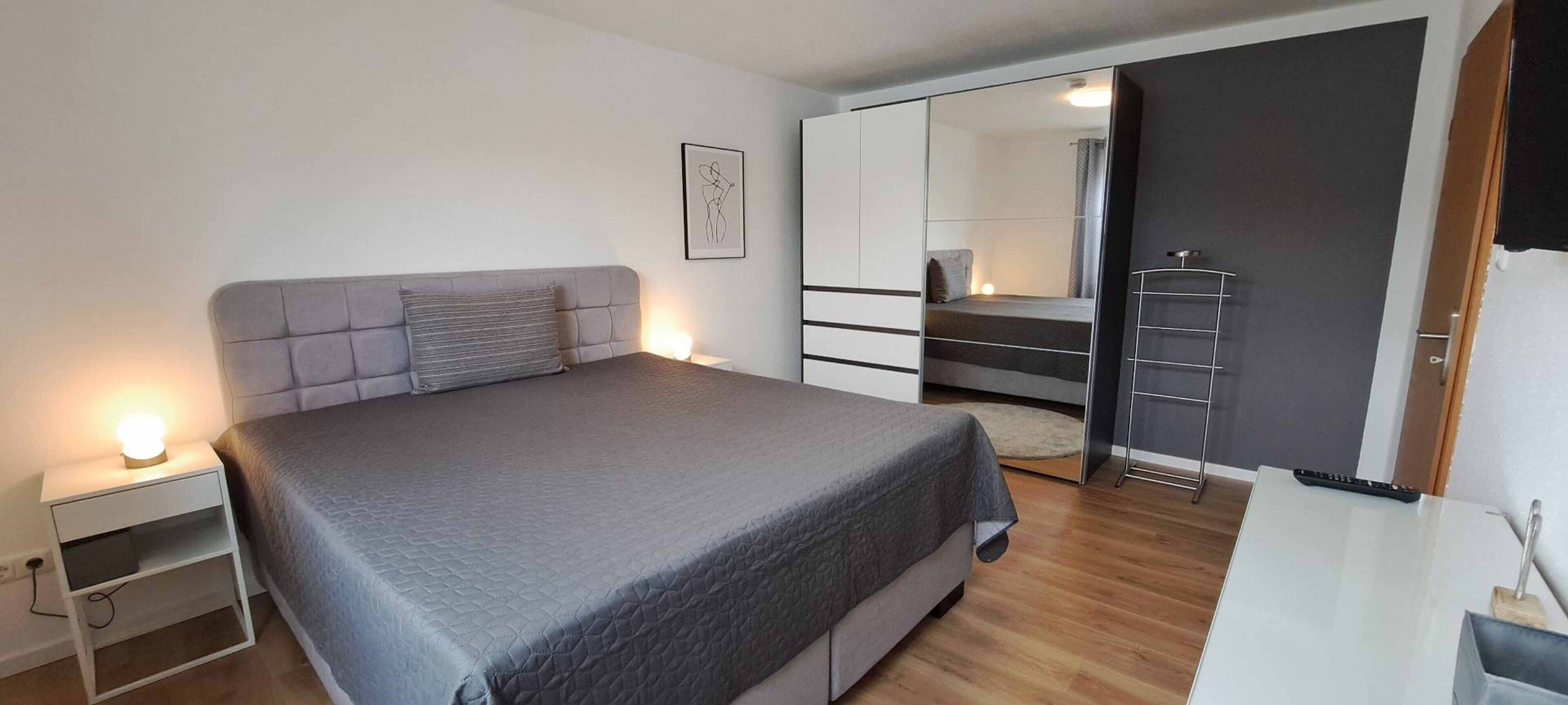A modern bedroom with a large bed, bedside tables, a wardrobe, and a mirror.