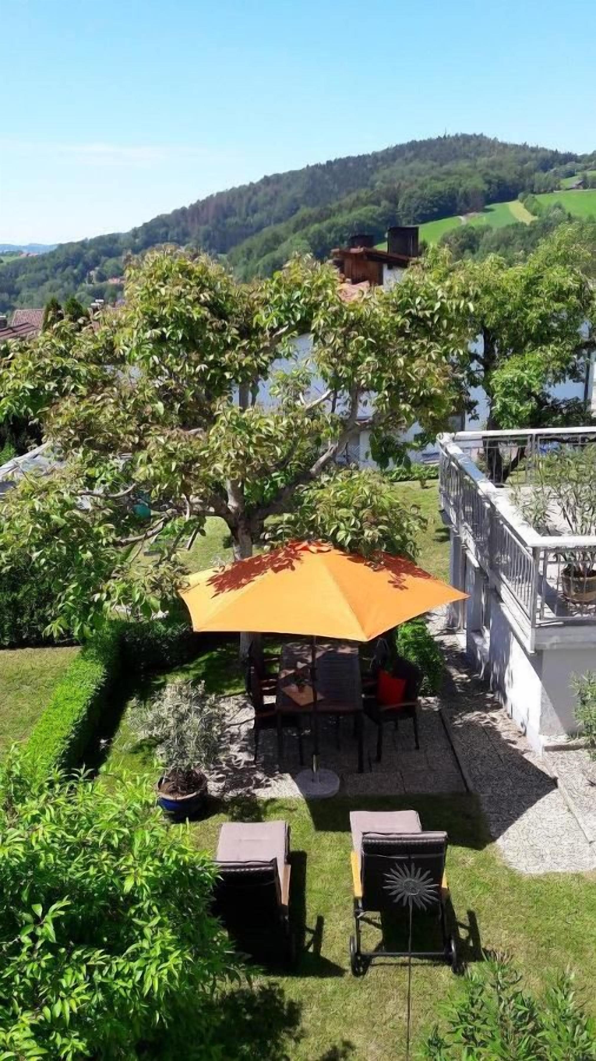 A patio area with a table and chairs under a large umbrella, surrounded by greenery and overlooking a scenic landscape.