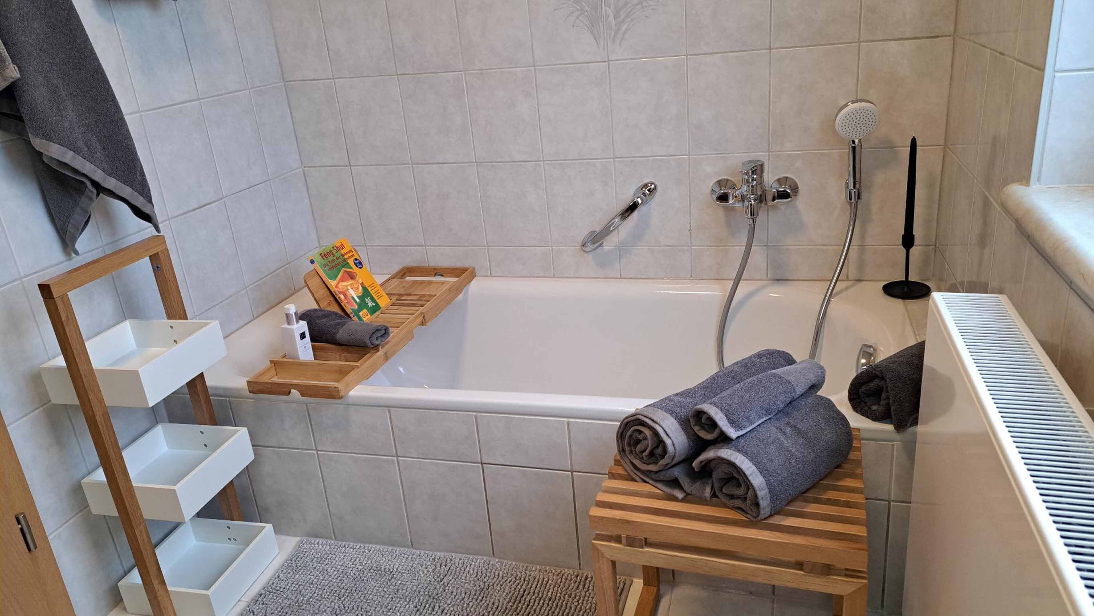 Et badeværelse med badekar, bruser, håndklædestige og en træhylde med toiletartikler.