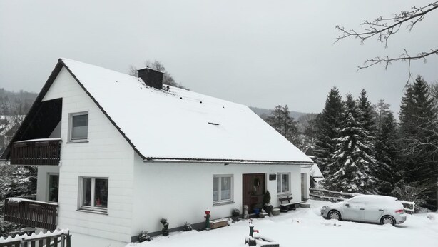 Une maison blanche à deux étages, dotée d’un toit à forte pente, entourée d’arbres couverts de neige et avec une voiture garée devant.
