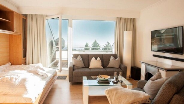 A hotel room with a bed, a sofa, a coffee table, a TV, and a balcony with a view. - Ferienwohnung Frohwein (Winterberg)