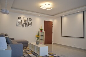 A modern living room with a grey sofa, a white coffee table, and a patterned rug. There is a projector mounted on the ceiling, a white shelf with decorative items, and a door in the background. - Beautiful 2-bedroom apartment - 63 m² - 50 m from the RER A station (Bussy-Saint-Georges)