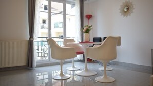 A modern living room with a balcony, a dining area with two chairs, a red chair, a small table with a vase of flowers, and a decorative wall piece. - Beautiful F3 apartment - 63m2 - 50m from RER A station (Bussy-Saint-Georges)