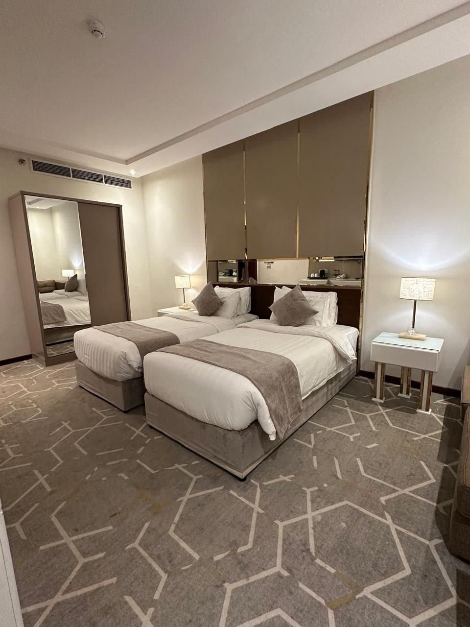 A hotel room with two beds, a large mirror, a bedside table with a lamp, and a patterned carpet.