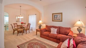 A living room with a red sofa, two lamps, a rug, and a dining area with chairs and a table. - South-Facing Private Pool • 2 Ensuites • 15 Minutes to Disney (Davenport)