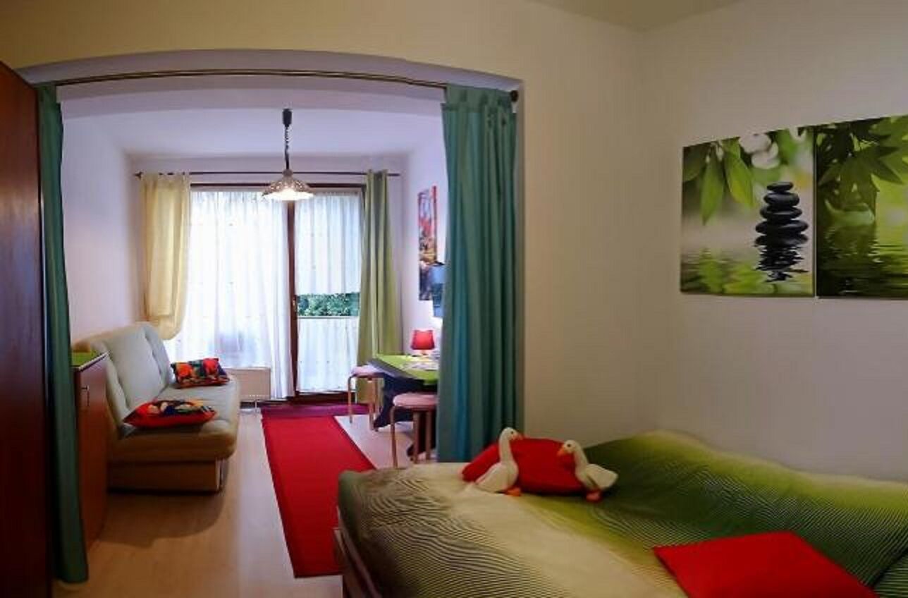 A hotel room with a bed, a sofa, a desk, and a chair. There is a window with curtains, a painting on the wall, and a red rug on the floor.