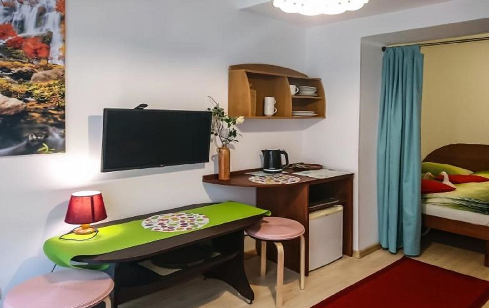 Una habitación funcional con escritorio, una silla, una cama, televisión y un pequeño estante con tazas y platos.