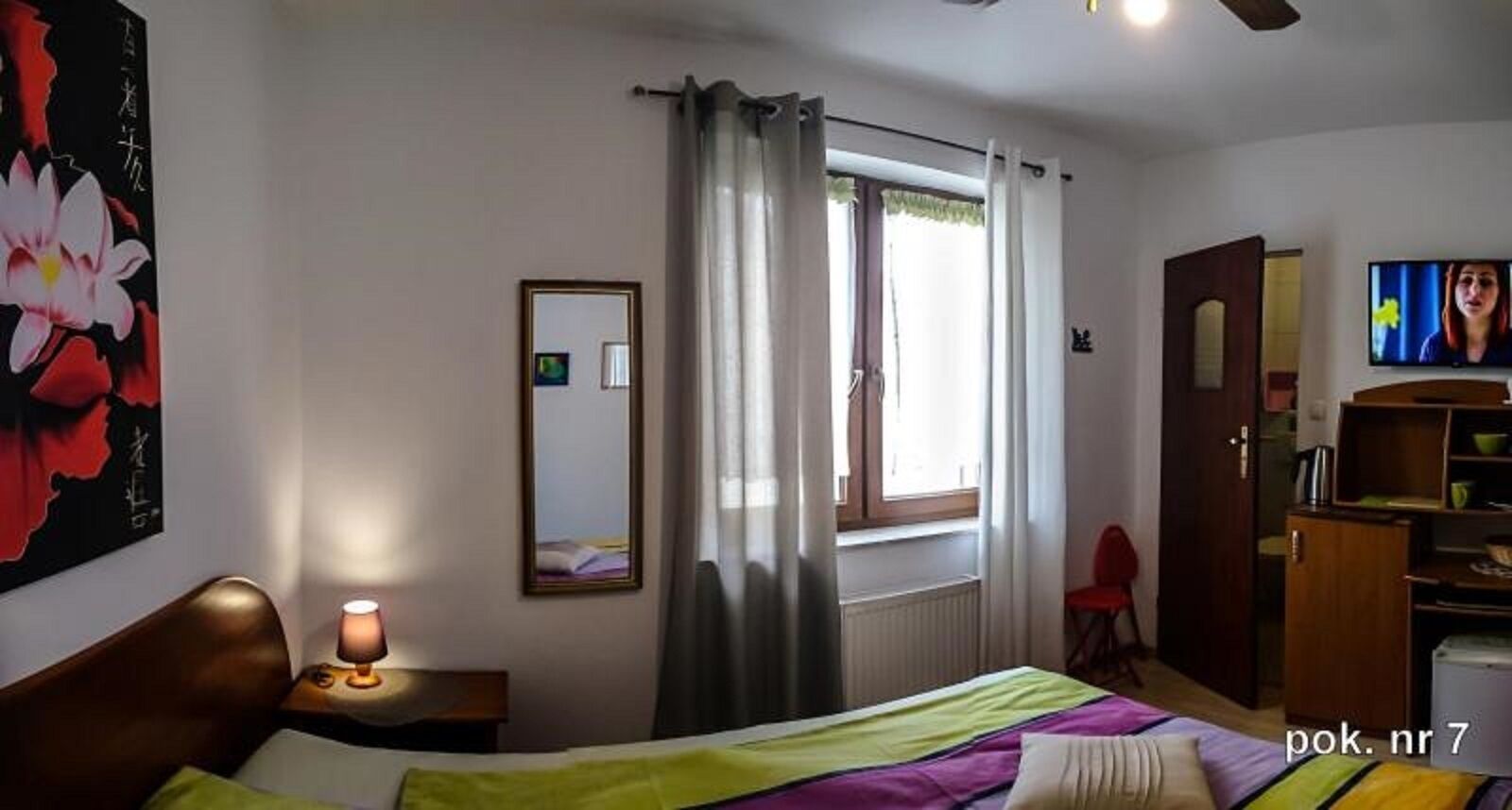 A bedroom with a bed, a bedside lamp, a window with curtains, a mirror, a television, and a wardrobe.