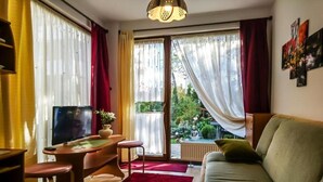 A living room with a TV, a sofa, a small table, and a chair. There are curtains on the windows, and a view of greenery outside. - Willa Christo Dom z Dusza (Kolobrzeg)