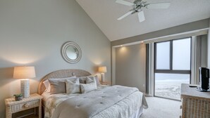 Un dormitorio con una cama grande, dos mesitas de noche con lámparas, un espejo y vistas a la playa a través de una puerta corrediza.