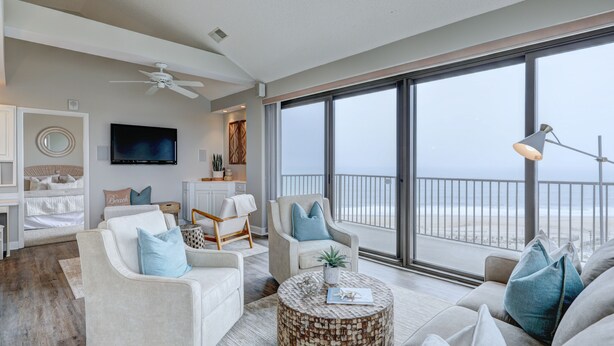 Un amplio salón con un gran ventanal con vista a una playa, un ventilador de techo y un televisor de pantalla plana montado en la pared.